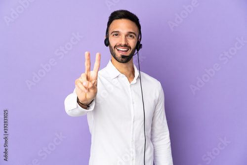 Telemarketer man working with a headset isolated on purple background smiling and showing victory sign © luismolinero