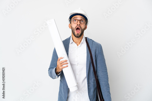 Young architect man with helmet and holding blueprints isolated on white background looking up and with surprised expression