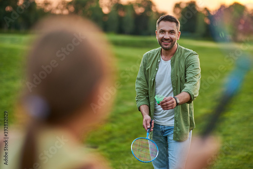 Happy father holding badminton racket and shuttlecock while playing with his little daughter outdoors in the park on a summer day © Kostiantyn