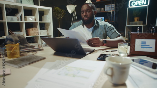Portrait of focused african man reading documents near laptop in night office.