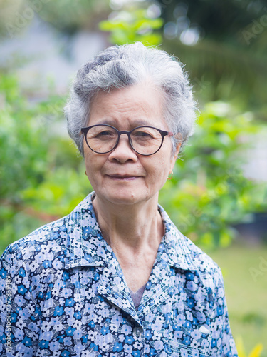 Elderly Asian woman wearing glasses, smiling and looking at the camera while standing in a garden. Concept of aged people and healthcare