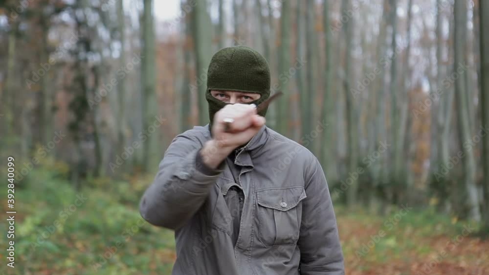 A man wearing a balaclava mask brandishes a large knife while standing in the woods. A man with a covered face threatens with a knife. A man wearing a balaclava mask brandishes a large knife while standing in the woods. A man with a covered face threatens with a knife.