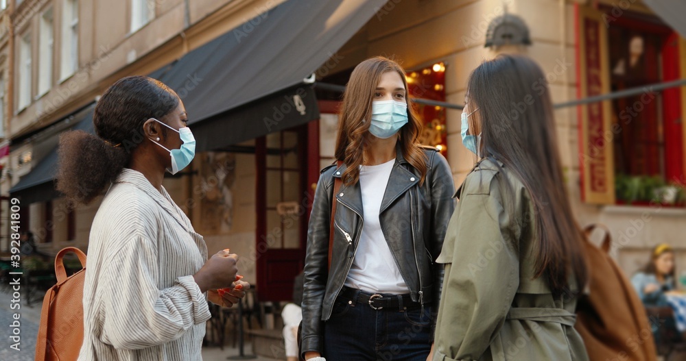 Three mixed-races young women, best friends meeting at city street and talking joyfully during coronavirus pandemic. Multi ethnic females in medical masks speaking and discussing something outdoor.