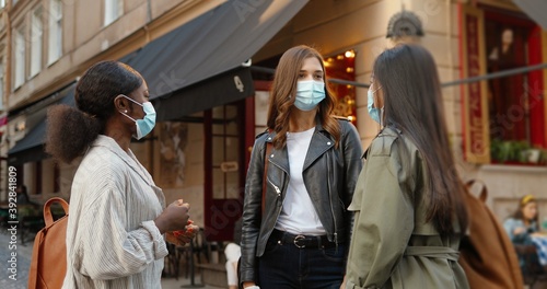Three mixed-races young women, best friends meeting at city street and talking joyfully during coronavirus pandemic. Multi ethnic females in medical masks speaking and discussing something outdoor.