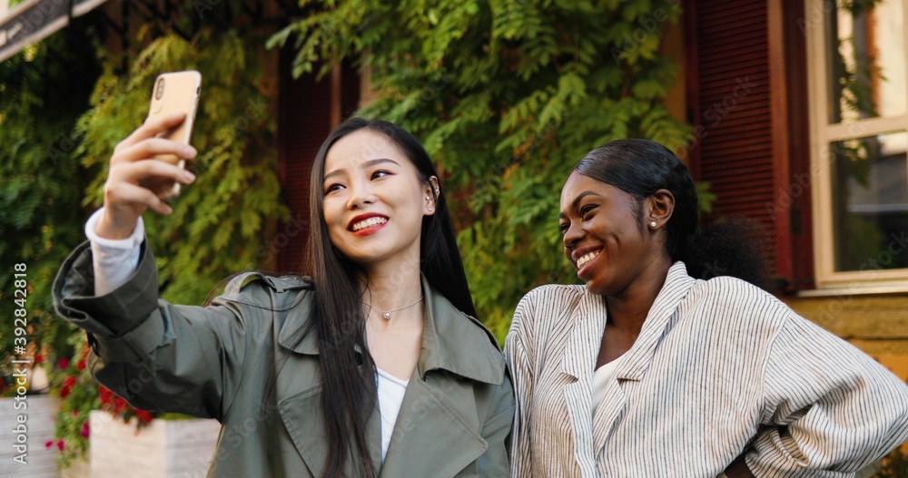 Cheerful Asian and African American beautiful females posing and smiling to smartphone camera. Mixed-races young women taking selfie photo with phone. Multi ethnic girls making photos outdoors. Cheerful Asian and African American beautiful females posing and smiling to smartphone camera. Mixed-races young women taking selfie photo with phone. Multi ethnic girls making photos outdoors.