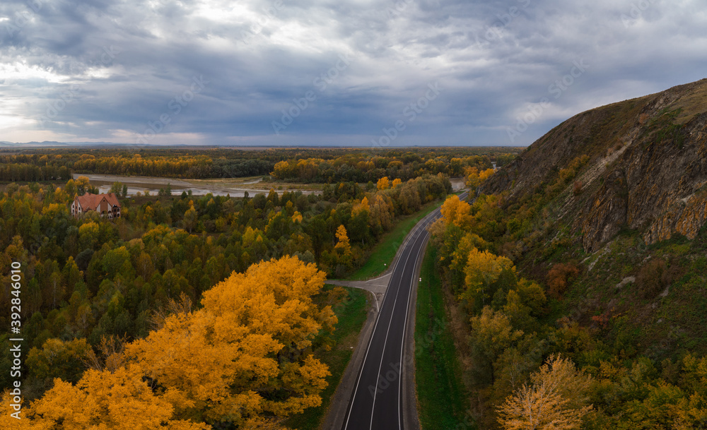 Aerial view of road in beautiful autumn Altai forest. Beautiful landscape with empty rural road, golden autumn in altai: trees with red, yellow and orange leaves.