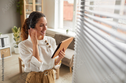 High angle portrait of smiling businesswoman wearing headset and holding digital tablet during online meeting in office, copy space