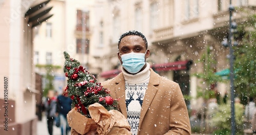 Close up portrait of joyful African American man in medical mask standing on street in snowy town. Handsome happy male holding little new year tree outdoors. Christmas concept.