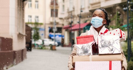 Beautiful African American woman walking in decorated city with many christmas gift boxes after xmas shopping. Portrait of joyful grateful female in mask holds holiday presents. Surprise gifts concept