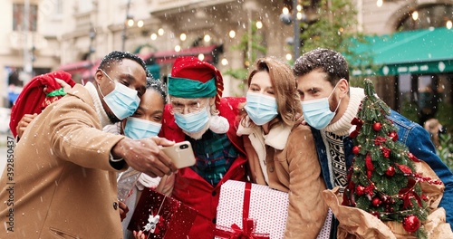 Santa Claus in mask hugs African American and Caucasian young people while taking selfie photo on cellphone. Portrait of males ad females friends take pictures with Santa on snowy street. X-mas spirit