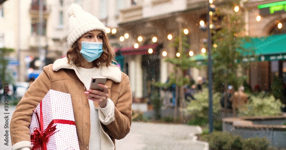 Portrait of young Caucasian beautiful female in mask standing in snowy decorated crowded city and texting on cellphone. Woman with christmas present tapping on smartphone. Winter holidays concept
