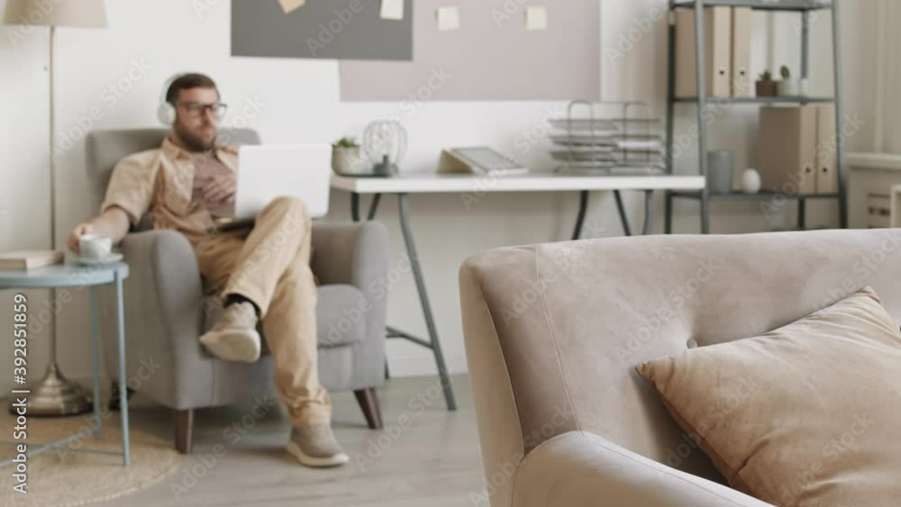 Full shot of young Caucasian man wearing over ear headphones sitting with legs crossed in armchair with laptop, drinking from coffee cup, nodding, talking. Guy having video call at home Full shot of young Caucasian man wearing over ear headphones sitting with legs crossed in armchair with laptop, drinking from coffee cup, nodding, talking. Guy having video call at home