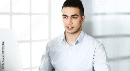 Businessman is working with computer in modern office. Headshot of arab male entrepreneur at workplace. Business concept