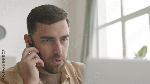Close-up of male brown-haired Caucasian worker wearing headset in one-ear, talking, nodding, saying goodbye, looking at laptop. Man having business video call at home