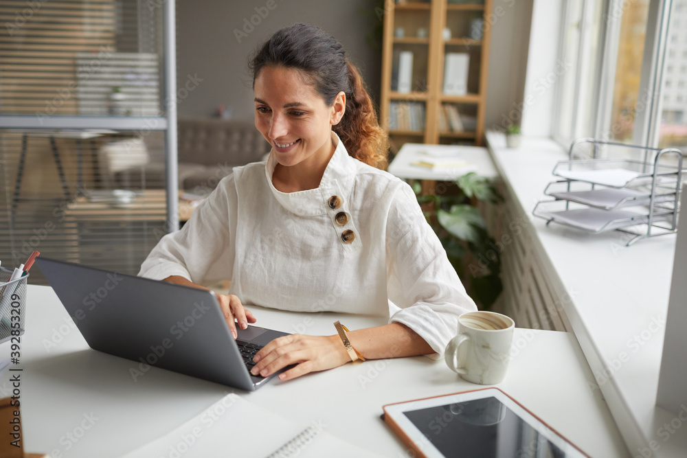 Portrait of cheerful adult businesswoman using laptop and smiling while working at desk in modern home office interior, copy space Portrait of cheerful adult businesswoman using laptop and smiling while working at desk in modern home office interior, copy space