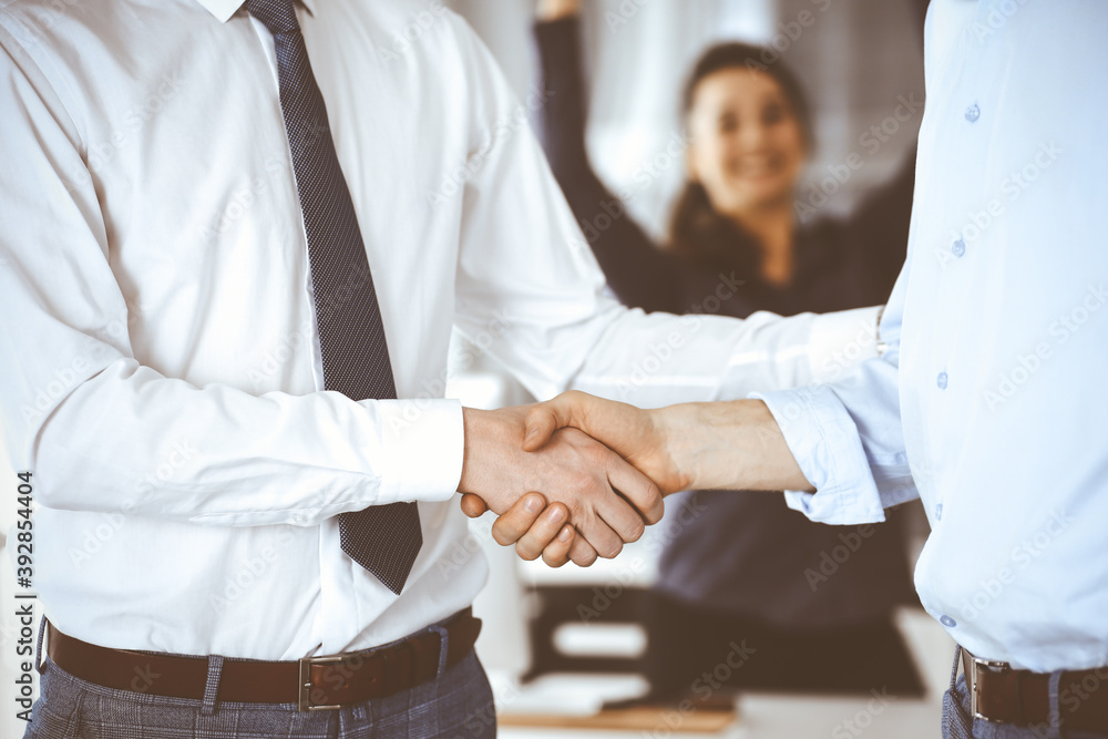 Fototapeta premium Two businessmen are shaking hands in office, close-up. Happy and excited business woman stands with raising hands at the background. Business people concept