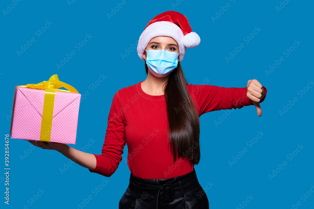 Obraz premium Young woman wearing Santa hat wearing face protective medical mask holding christmas or new year gift box and showing thumb down gesture, blue isolated background.