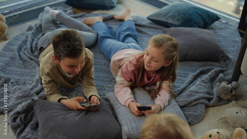 Kids gaming smartphones. Siblings lying with cell phones next kid on carpet.