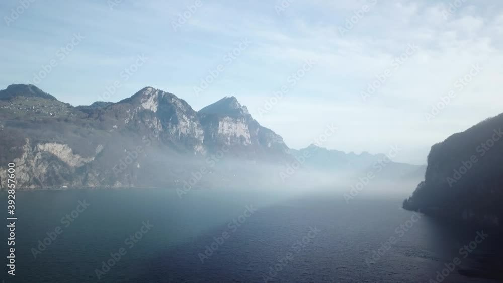 Mysterious mystical atmosphere created by foggy clouds hanging between two mountains. Panoramic shot of mountain ranges above crystal blue mountain lake. Mysterious mystical atmosphere created by foggy clouds hanging between two mountains. Panoramic shot of mountain ranges above crystal blue mountain lake.