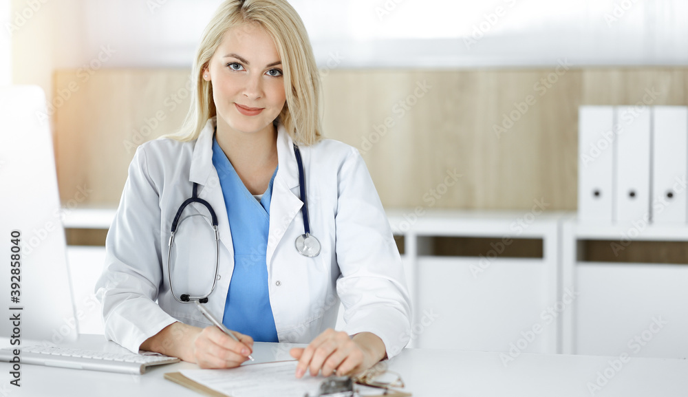 Woman-doctor at work while sitting at the desk in sunny clinic. Blonde cheerful physician filling up medication history record form Woman-doctor at work while sitting at the desk in sunny clinic. Blonde cheerful physician filling up medication history record form