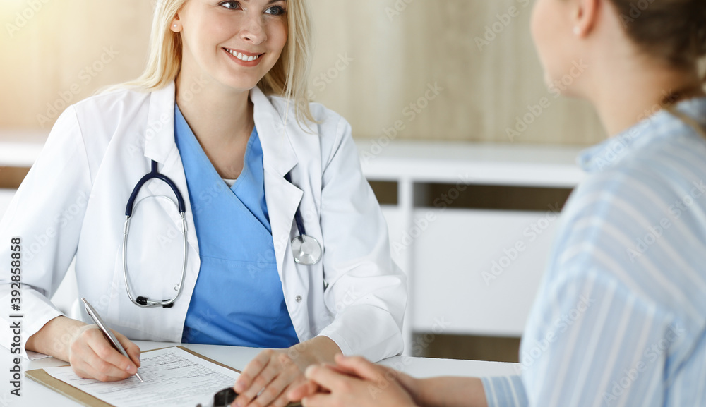 Fototapeta premium Woman-doctor and patient sitting and talking in sunny clinic. Blonde therapist is cheerfully smiling