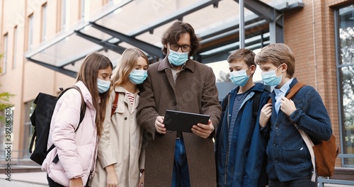 Portrait of Caucasian handsome happy male professor in glasses and mask tapping on tablet outdoors. Teacher with girls and boys school students browsing on device near school. Education concept