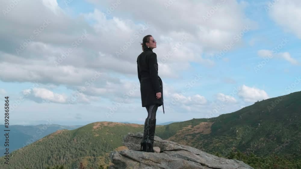 beautiful girl in black cloak stands on a rock in high-heeled boots and sunglasses, against a background of mountains, view from the back, turning head admiring the nature. clear day, sky with clouds beautiful girl in black cloak stands on a rock in high-heeled boots and sunglasses, against a background of mountains, view from the back, turning head admiring the nature. clear day, sky with clouds