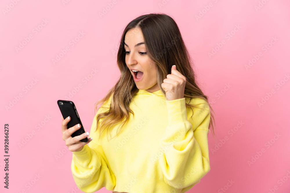 Young Romanian woman isolated on pink background using mobile phone and doing victory gesture Young Romanian woman isolated on pink background using mobile phone and doing victory gesture