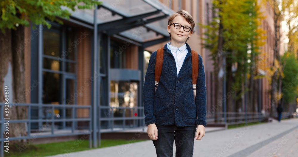Portrait of joyful Caucasian boy pupil in glasses in good mood standing outdoor near school. Happy male student with backpack smiling to camera on street in town. Education concept Portrait of joyful Caucasian boy pupil in glasses in good mood standing outdoor near school. Happy male student with backpack smiling to camera on street in town. Education concept