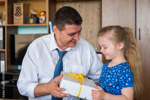 A little girl in a blue dress gives her dad a white box with a gift.