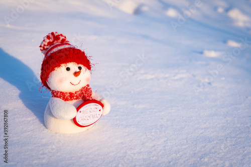 A toy snowman in a red hat on the snow in the rays of light wishes you a merry Christmas