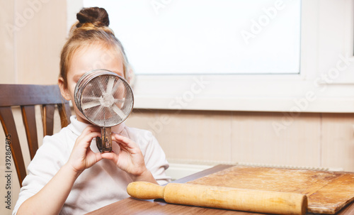 cute preschoolgirl girl holding flour sifter and looks through it. cooking concept. on the kitchen photo