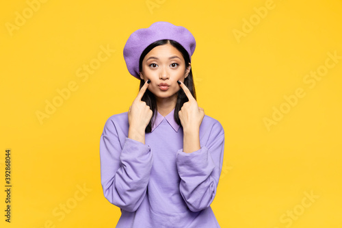Canvas Print asian woman in beret touching cheeks with fingers while pouting lips isolated on