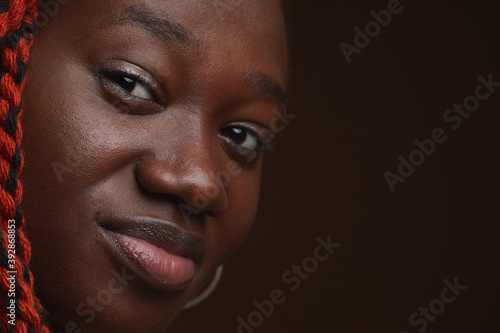 Extreme close up portrait of young African-American woman with braided hair looking at camera while posing in against dark brown background in studio, copy space
