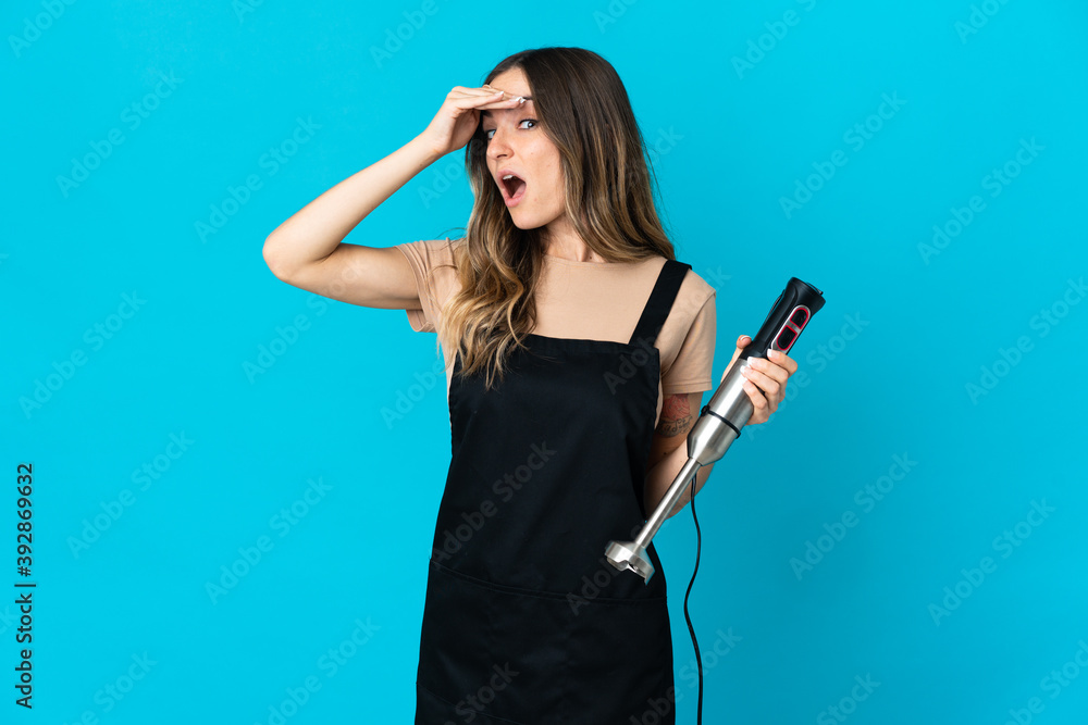 Romanian woman using hand blender isolated on blue background doing surprise gesture while looking to the side Romanian woman using hand blender isolated on blue background doing surprise gesture while looking to the side