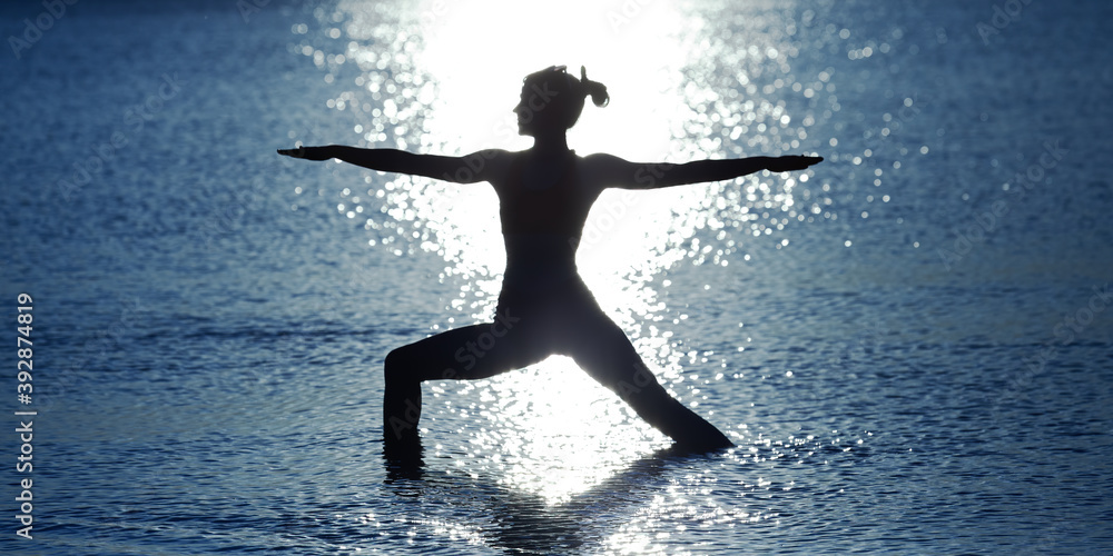Silhouette of a slender girl practicing yoga in the water. Background of a blue sea (ocean, lake) and sun reflections. Standing position of yoga as exercise - asana  The Warrior (Virabhadrasana).