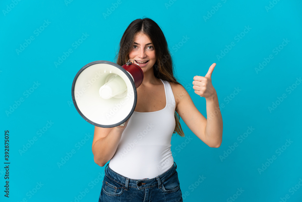 Young caucasian woman isolated on blue background shouting through a megaphone to announce something and with thumb up Young caucasian woman isolated on blue background shouting through a megaphone to announce something and with thumb up