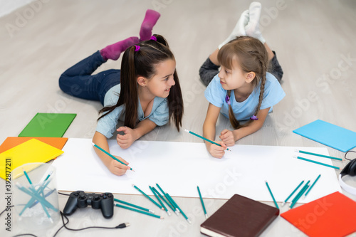 two little girls sisters lie on the floor of the house and draw with colored pencils on paper. children do creative homework after school.