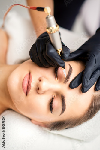 Beautician applying permanent makeup on eyebrows of young woman by special tattoo machine tool