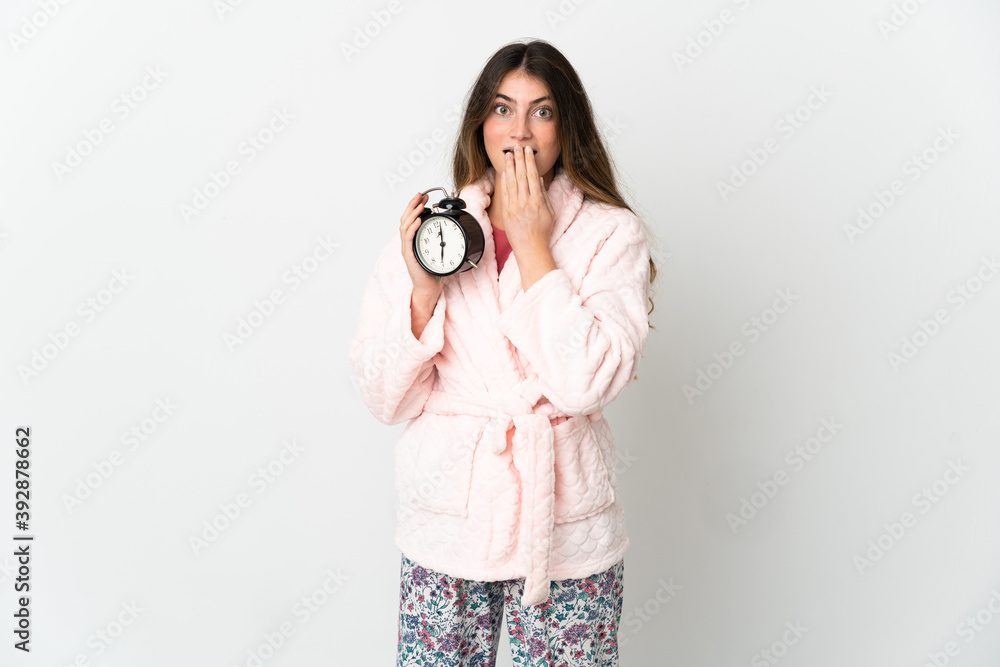 Young caucasian woman isolated on white background in pajamas and holding clock with surprised expression Young caucasian woman isolated on white background in pajamas and holding clock with surprised expression