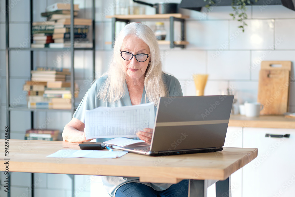 Senior woman using laptop for websurfing in her kitchen. The concept of senior employment, social security. Mature lady sitting at work typing a notebook computer in an home office. Senior woman using laptop for websurfing in her kitchen. The concept of senior employment, social security. Mature lady sitting at work typing a notebook computer in an home office.