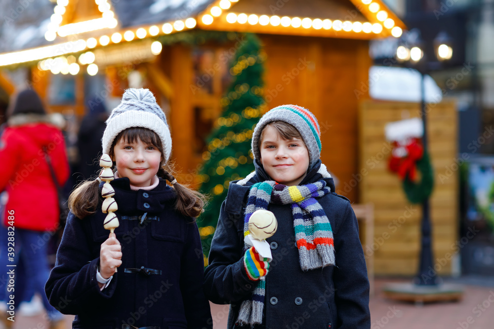 Little cute kid girl and boy eating white chocolate covered strawberries and apple on skewer on traditional German Christmas market. Happy children, best friends, twins and siblings on snowy day