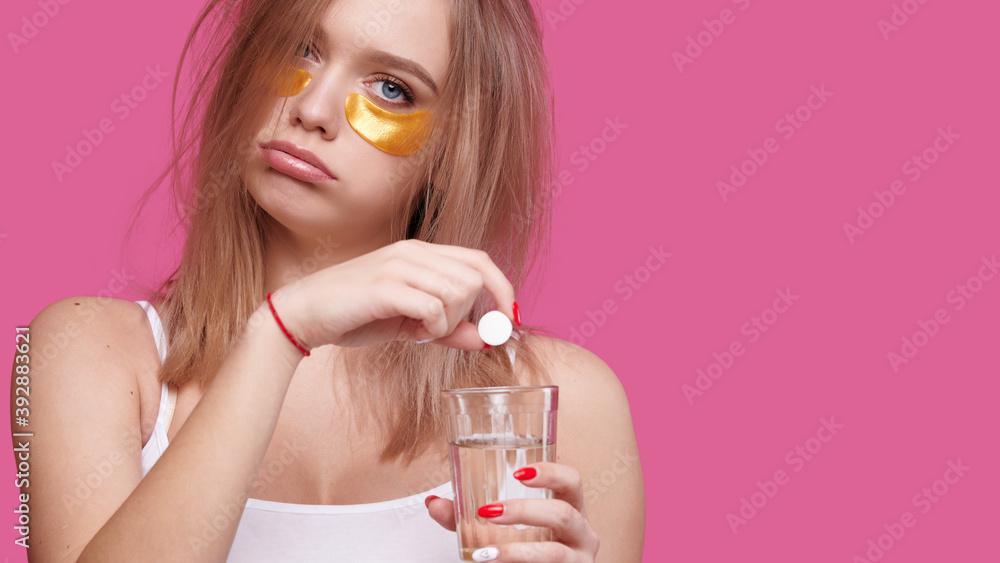 Sleepy young woman with patches under the eyes puts pain relieving tablet into glass of water. Girl after hangover early in the morning, pink isolated background Sleepy young woman with patches under the eyes puts pain relieving tablet into glass of water. Girl after hangover early in the morning, pink isolated background