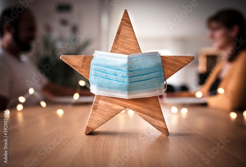 Wooden star with a face mask and a couple in the background, celebrating Christmas alone at home photo