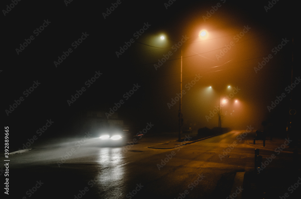 one moving car in a foggy city road at night with headlights on