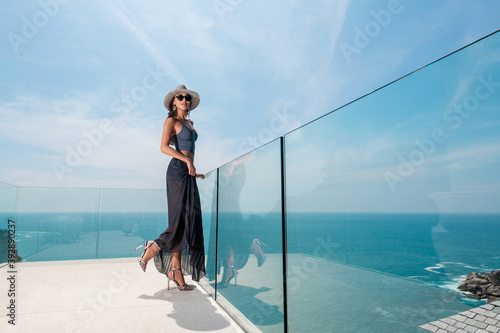 Fashionable girl with stylish accessories in a hat, a long skirt posing on a transparent balcony overlooking the sea and rainforest