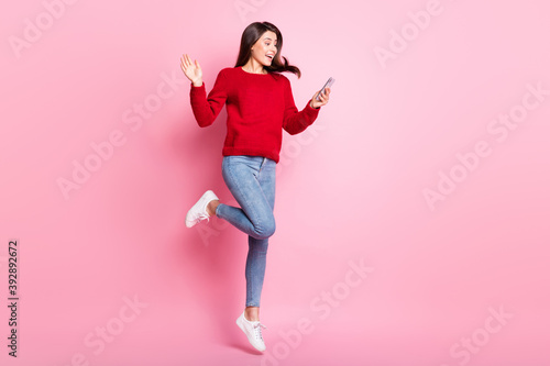 Full length photo of pretty girl waving hand screen hold cellphone jump wear red pullover jeans sneakers isolated pink color background