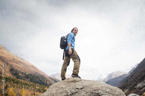 An adult man with a backpack stands on a rock, he travels through the mountains. Autumn landscape in the gorge, He stands against the sky and looks from above. Karachay Cherkessia, Russia.