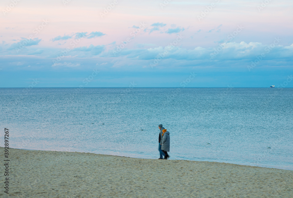 Elderly couple is walking on the beach at sunset on a beautiful autumn day. Colorful pink blue sky and calm sea with ship in the background.