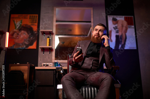 bearded macho man, barber male indoors, confident caucasian barber owner sits on chair posing, holding electric razor in hands